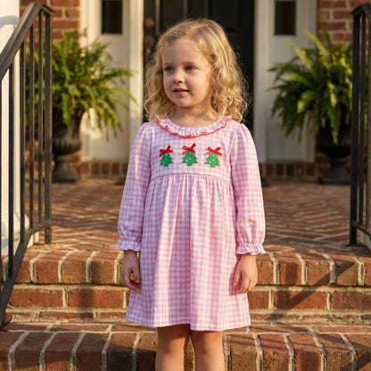 A young girl is wearing a pink and white gingham check dress with long sleeves and a ruffled collar. The bodice features smocking detailed with three embroidered green Christmas trees topped with red bows.