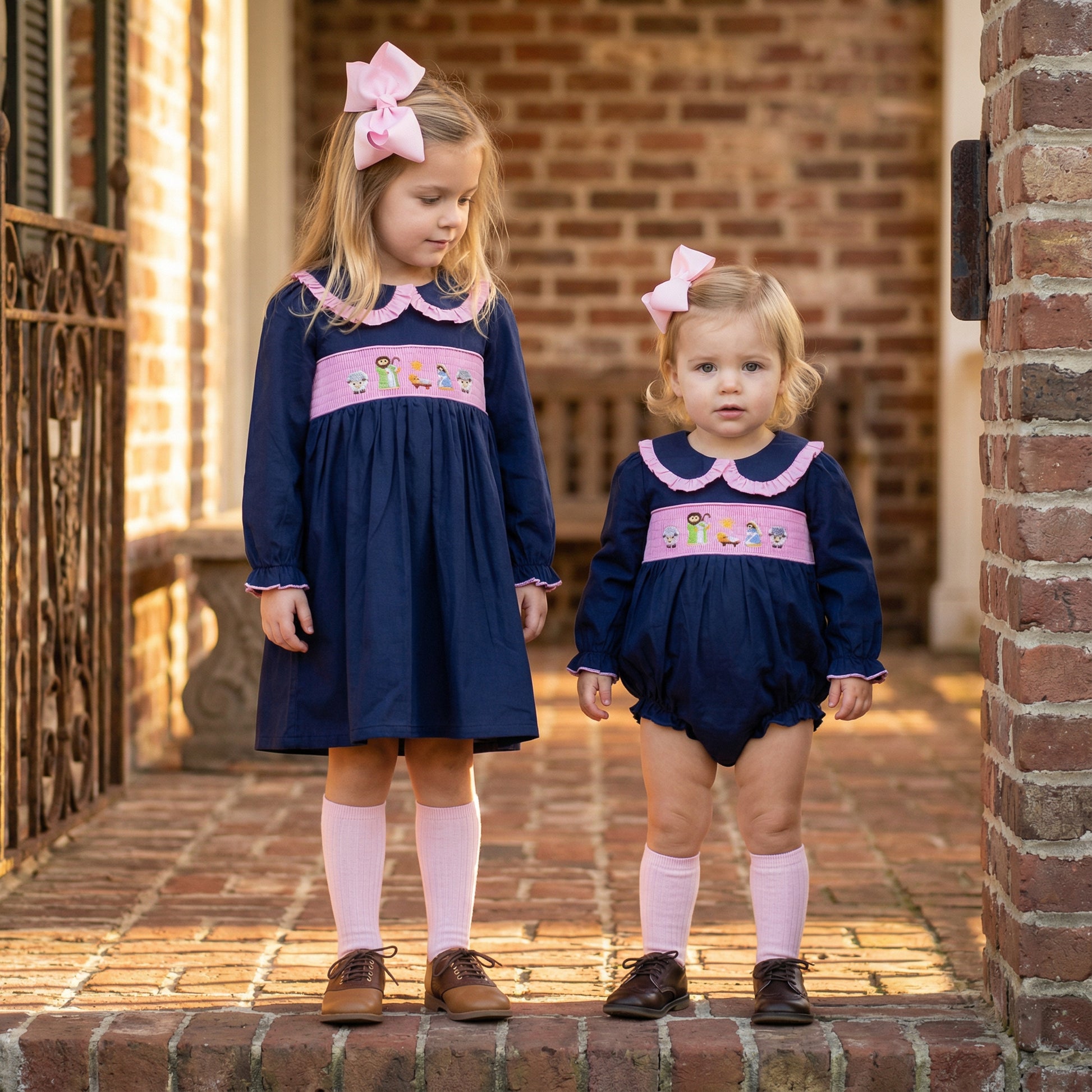 Two young girls stand side-by-side on a brick porch, wearing matching navy blue outfits with pink accents. The older girl on the left wears a long-sleeved dress with a pink ruffled collar and a pink smocked panel across the chest embroidered with a Nativity scene featuring Mary, Joseph, Jesus, and sheep. She wears pink knee-high socks and brown saddle shoes. The younger toddler on the right wears a matching navy blue bubble romper with the same pink collar and Nativity smocking.