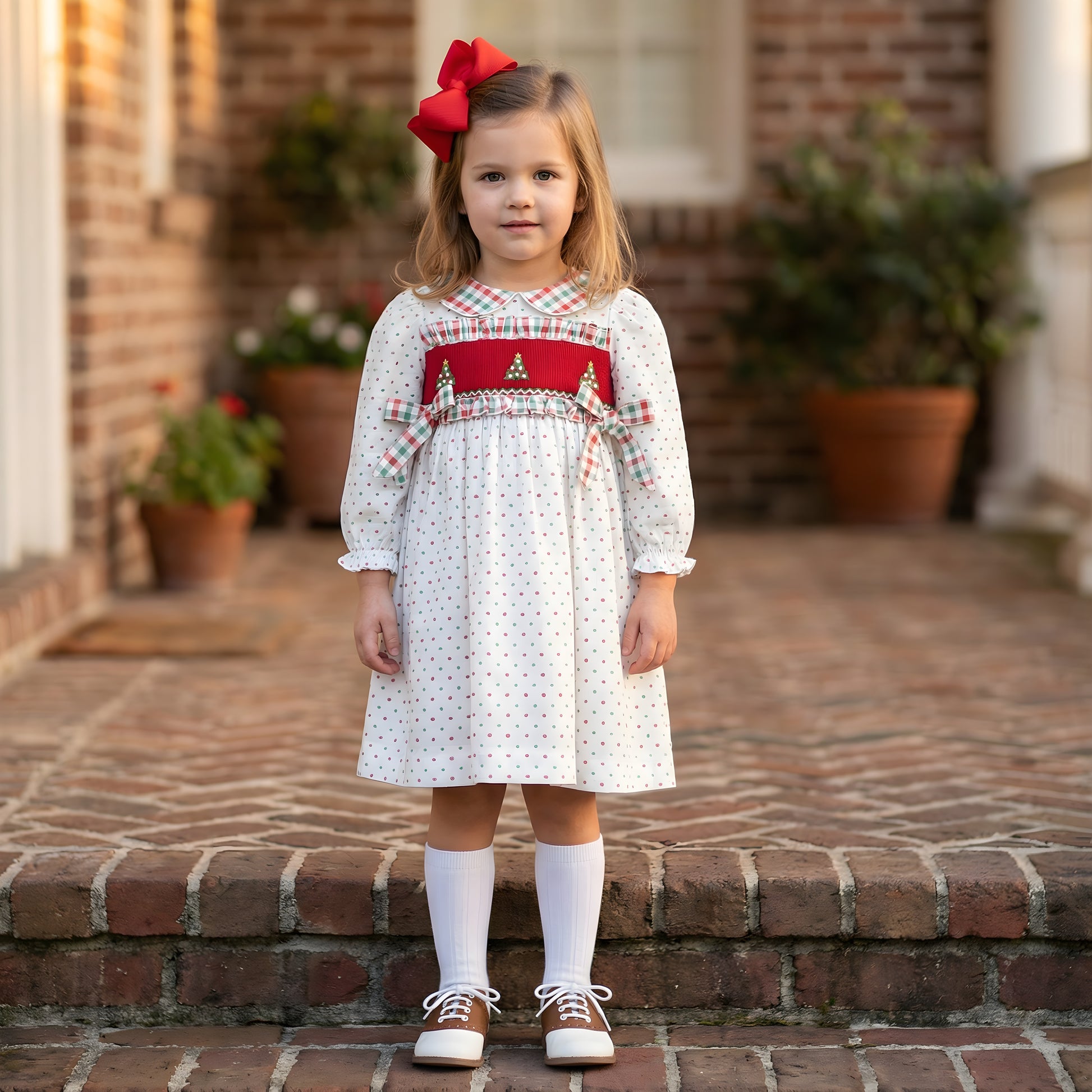 Young girl wearing a white Swiss Dot dress featuring a red hand smocked plate embroidered with green Christmas trees and accented with plaid ruffles and side bows, standing on brick steps.