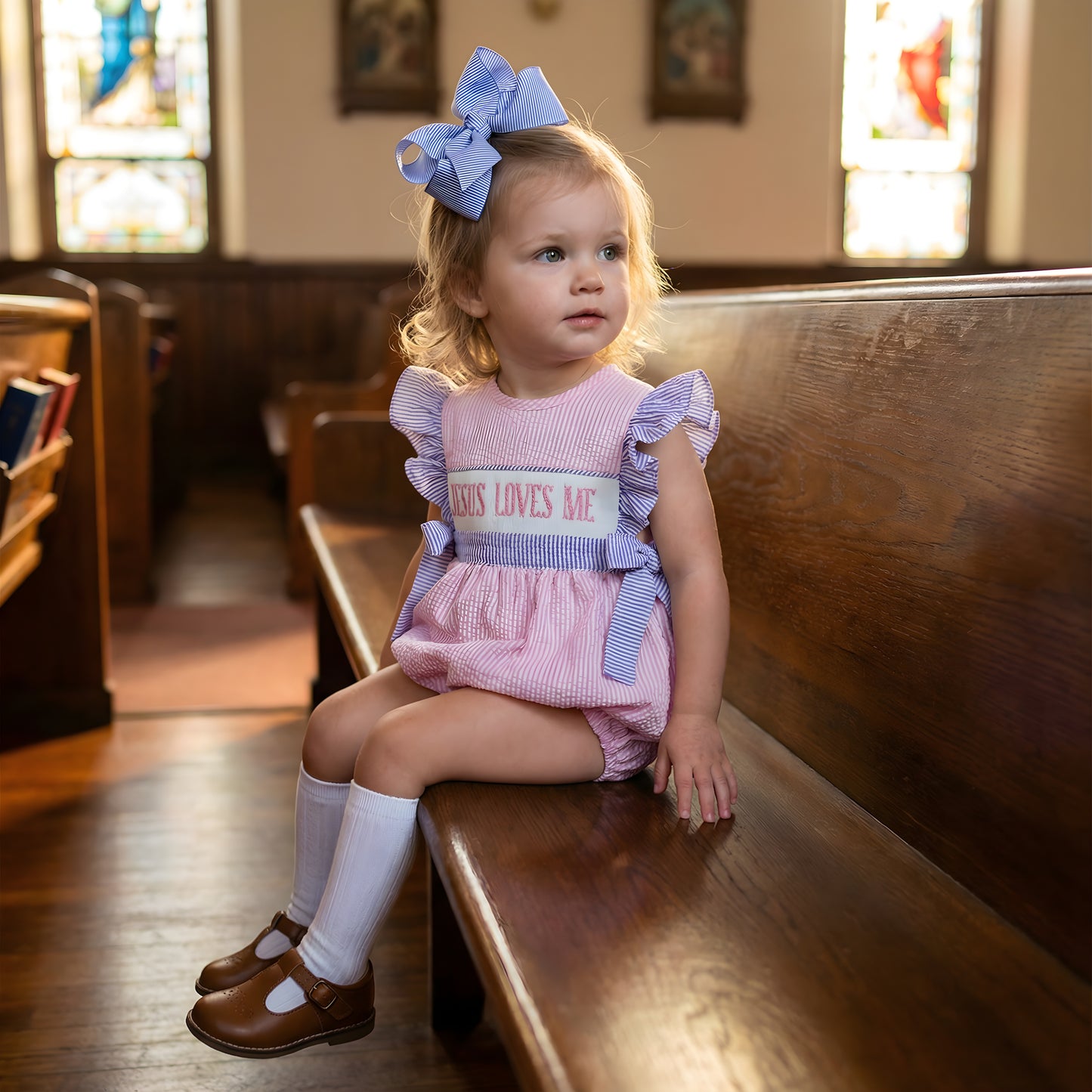 Toddler girl sitting on a church pew wearing a pink Hand Smocked Girl Bubble with Jesus Loves Me embroidery and a matching blue bow.