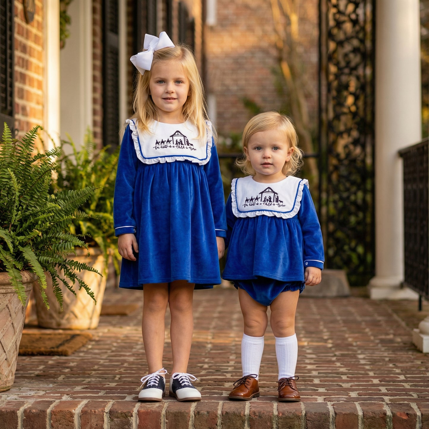 Two children standing on a brick walkway wearing matching royal blue holiday outfits. The older child is in a long-sleeve blue dress with a white bib-style collar featuring an embroidered nativity scene and the phrase ‘For Unto Us a Child is Born,’ paired with black sneakers and white socks. The younger child is in a blue diaper set with the same embroidered collar, paired with brown shoes and white knee-high socks.