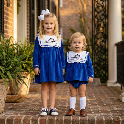 Two children standing on a brick walkway wearing matching royal blue holiday outfits. The older child is in a long-sleeve blue dress with a white bib-style collar featuring an embroidered nativity scene and the phrase ‘For Unto Us a Child is Born,’ paired with black sneakers and white socks. The younger child is in a blue diaper set with the same embroidered collar, paired with brown shoes and white knee-high socks.