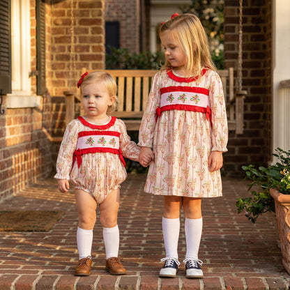 The image shows two young girls standing on a brick porch, holding hands and wearing matching outfits. The girl on the right wears a long-sleeved dress with a white fabric featuring a small pink floral pattern. It has red smocking across the chest with small embroidered details, a red ruffled collar, and red ribbons tied at the sides. She wears white knee-high socks and white and blue saddle shoes. The younger girl on the left wears a matching long-sleeved romper in the same floral fabric. 