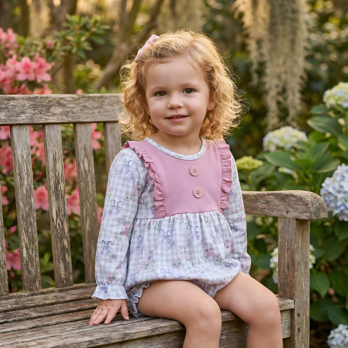 Toddler girl sitting on a bench wearing a Grandmillennial floral girls bow bubble romper featuring a grey gingham print with blue bows, pink flowers, and a ruffled solid pink bib.