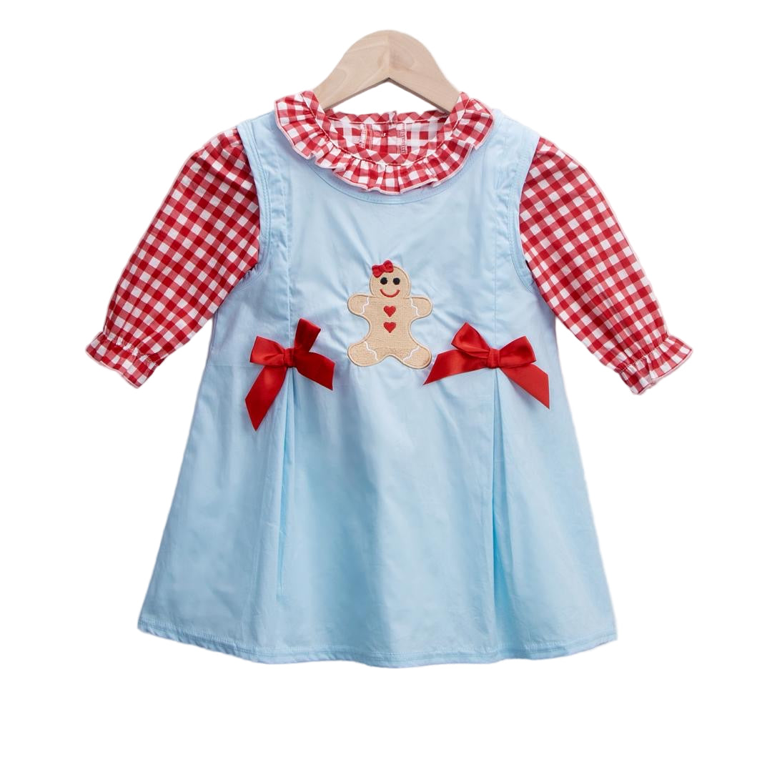 A festive baby girl outfit features a light blue jumper dress with a gingerbread girl applique and red bows, paired with a red and white gingham long-sleeve shirt with a ruffled collar, all hanging on a wooden hanger against a white background.