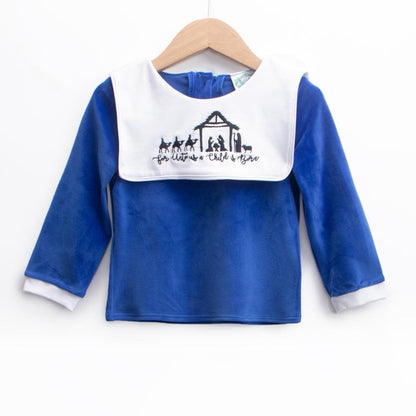 A royal blue long-sleeve top displayed on a wooden hanger. The top features white cuffs and a large white bib-style collar embroidered with a nativity scene and the phrase ‘For Unto Us a Child is Born.'