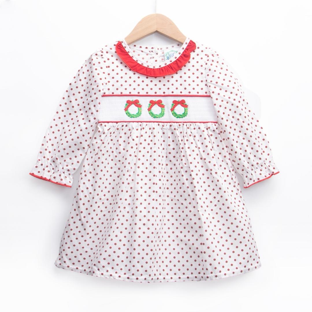 A white long-sleeved dress for a young girl featuring an all-over brown polka dot pattern, hanging on a wooden hanger. The bodice has a white smocked panel embroidered with three green Christmas wreaths topped with red bows. The neckline is accented with a red ruffled collar, and the sleeves feature matching red trim at the cuffs.