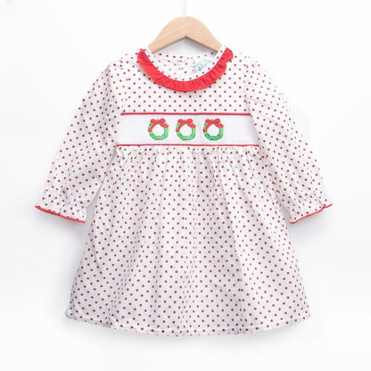 A white long-sleeved dress for a young girl featuring an all-over brown polka dot pattern, hanging on a wooden hanger. The bodice has a white smocked panel embroidered with three green Christmas wreaths topped with red bows. The neckline is accented with a red ruffled collar, and the sleeves feature matching red trim at the cuffs.