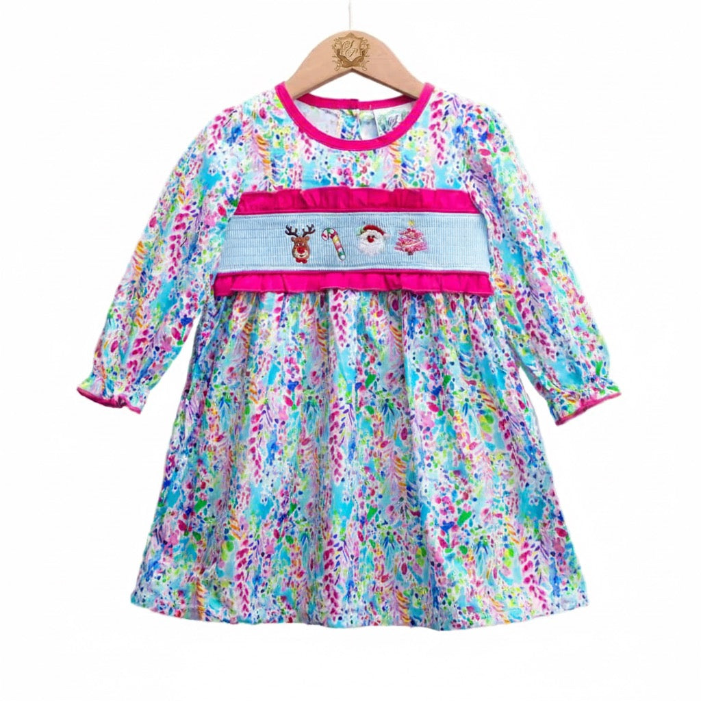 A child's long-sleeved dress with a colorful floral pattern in blue, pink, green, and white is hanging on a wooden hanger against a white background. The dress features a light blue smocked bodice with hot pink ruffles above and below it. Embroidered on the smocked panel are a reindeer, a candy cane, a Santa Claus face, and a decorated Christmas tree. The neckline and sleeve cuffs are also trimmed in hot pink. The wooden hanger has a logo printed on the center.