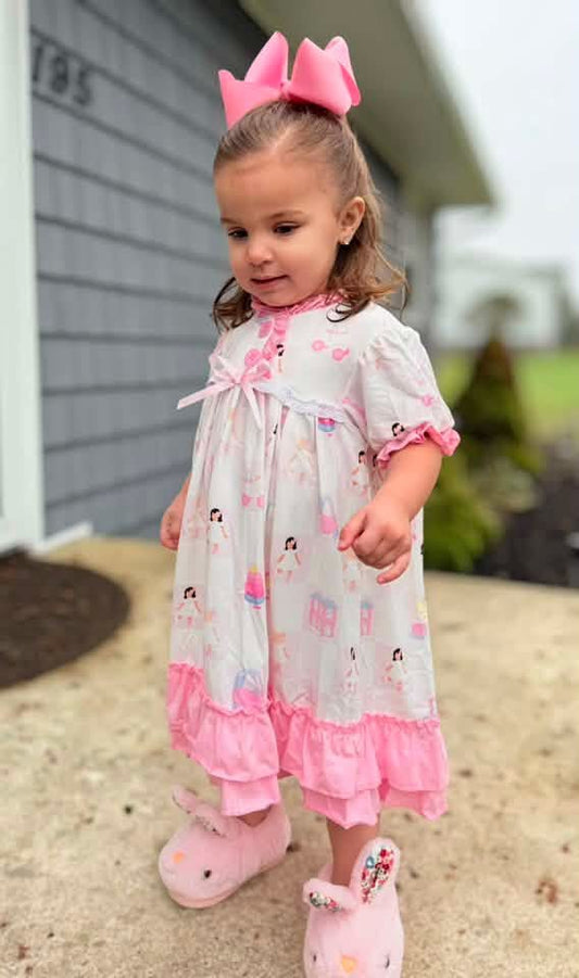 A toddler girl wearing a white and pink printed nightgown with pink ruffles, a pink bow headband, and pink slippers, standing outdoors in front of a house.