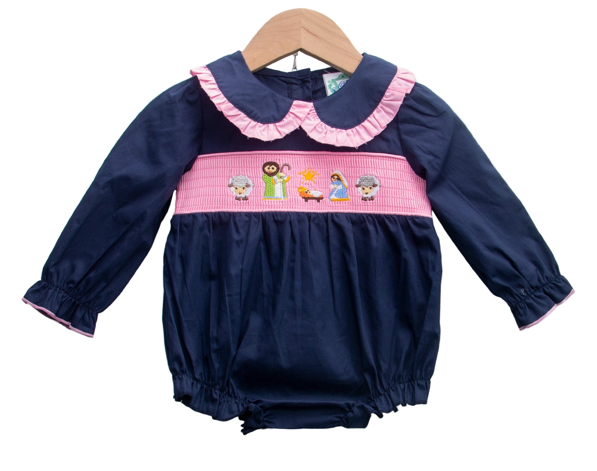 A baby romper displayed on a wooden hanger. The romper is navy blue with long sleeves and pink trim at the cuffs. It features a pink ruffled collar and a smocked waistband embroidered with nativity-themed characters, including figures and animals. The bottom has elastic leg openings.