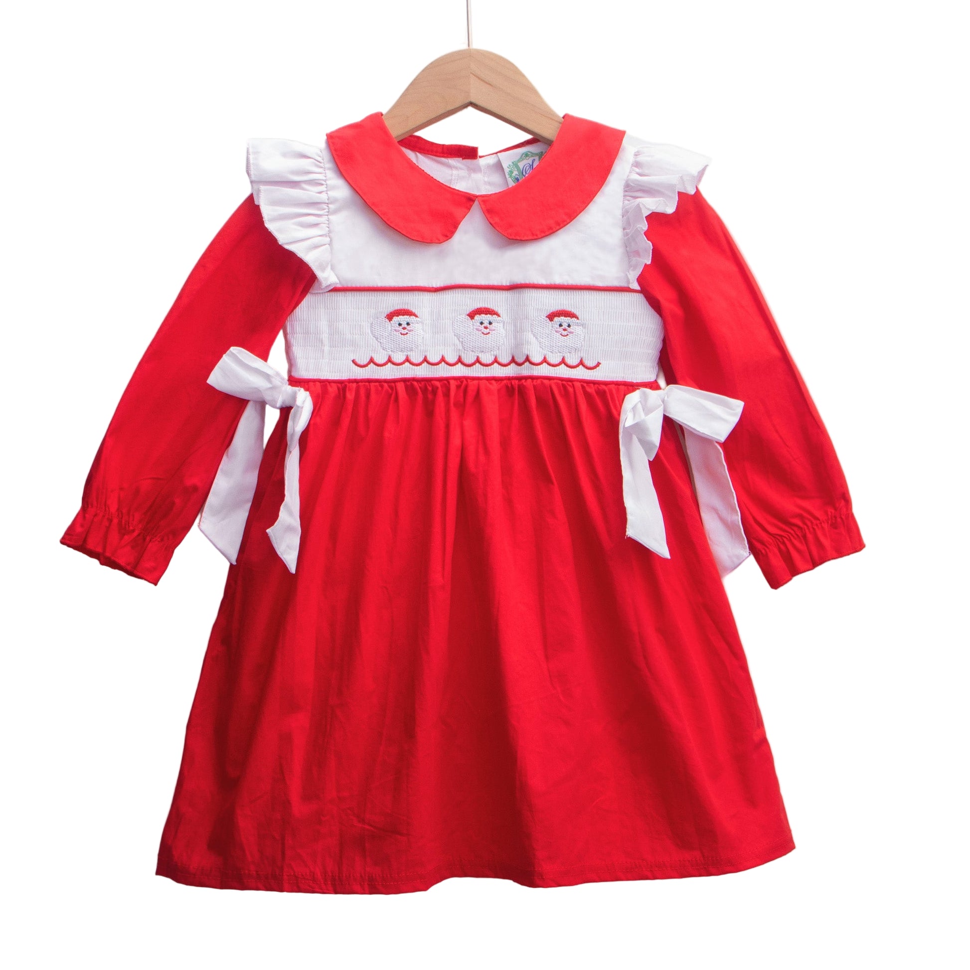 A red and white child's smocked dress hangs on a wooden hanger against a white background. The dress features a white Peter Pan collar with red trim, white ruffled shoulder accents, and a white smocked bodice with three embroidered Santa faces and a red wavy line below them. Long red sleeves end in elastic cuffs, and the red gathered skirt has a white bow tied at each side of the waist.