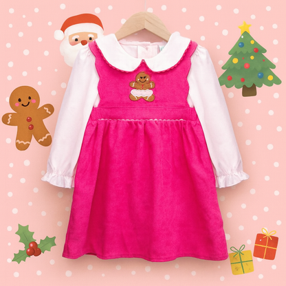 A studio photograph of a child's pink corduroy jumper dress layered over a white long-sleeved shirt with a Peter Pan collar, hanging on a wooden hanger against a pink background patterned with white polka dots and cartoon Christmas illustrations. The jumper features a small embroidered gingerbread man on the chest.