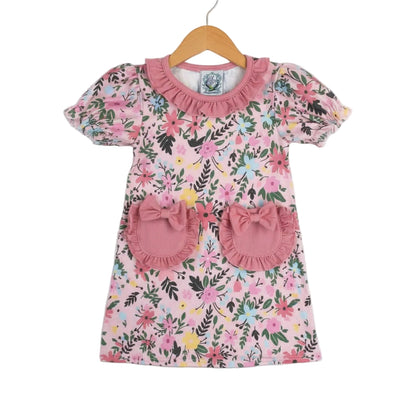 Front view of a girls' pink Field Of Flowers Spring Pocket Dress hanging on a wooden hanger, featuring a colorful floral print, puffed sleeves, a ruffled collar, and two front pockets adorned with pink ruffles and bows.