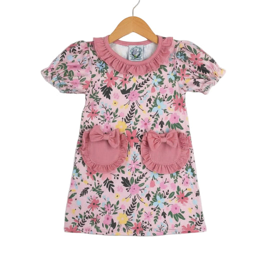 Front view of a girls' pink Field Of Flowers Spring Pocket Dress hanging on a wooden hanger, featuring a colorful floral print, puffed sleeves, a ruffled collar, and two front pockets adorned with pink ruffles and bows.