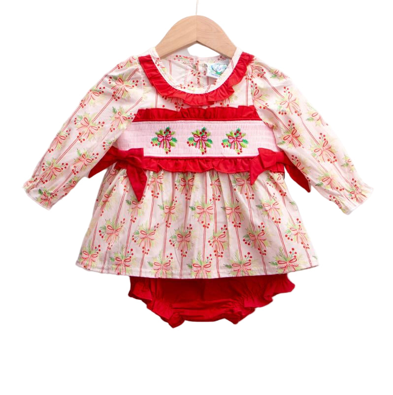 A baby diaper set displayed on a wooden hanger. The set includes a long-sleeve top with a white background and a repeating pink bow and green leaf pattern, accented by a red ruffled neckline and a smocked waistband featuring embroidered holiday bows with red ribbon details. Matching red diaper cover is visible underneath.
