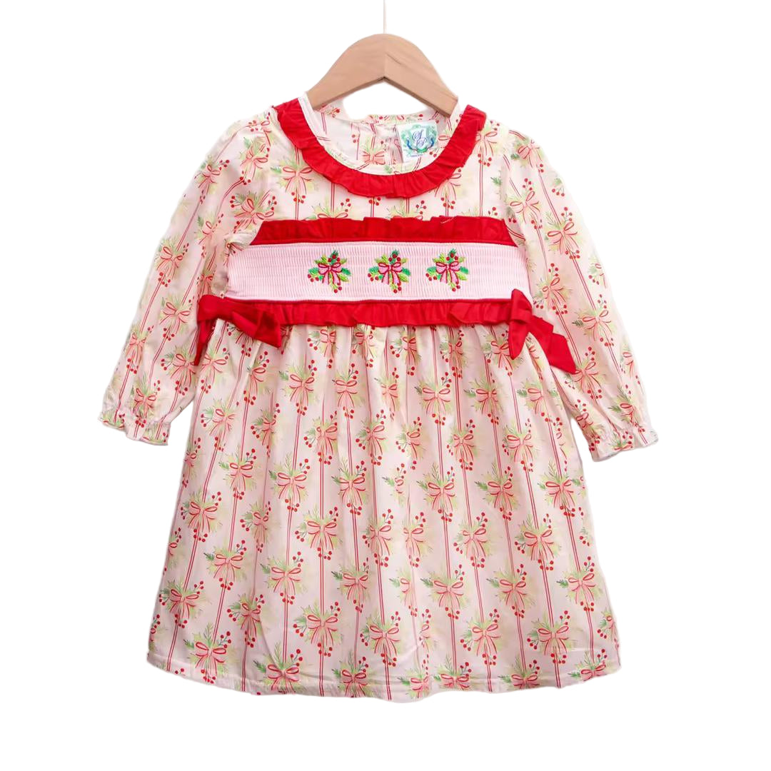 A long-sleeved child's dress on a wooden hanger. The light pink fabric is covered in a repeating pattern of red and green ribbons, bows, and holly sprigs. It has a red ruffled collar and matching red ruffles around the cuffs. Across the chest is a panel of white smocking with embroidered Christmas bows and holly in red and green thread. The dress hangs against a plain white background.