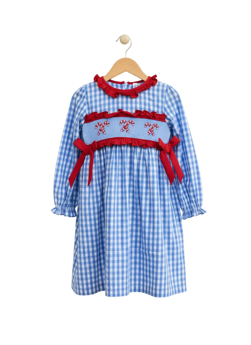 A girls' long-sleeved blue and white gingham dress hanging on a wooden hanger. It features a red ruffled collar and a smocked bodice embroidered with three pairs of candy canes, accented by red ruffles and red ribbon bows on the sides.