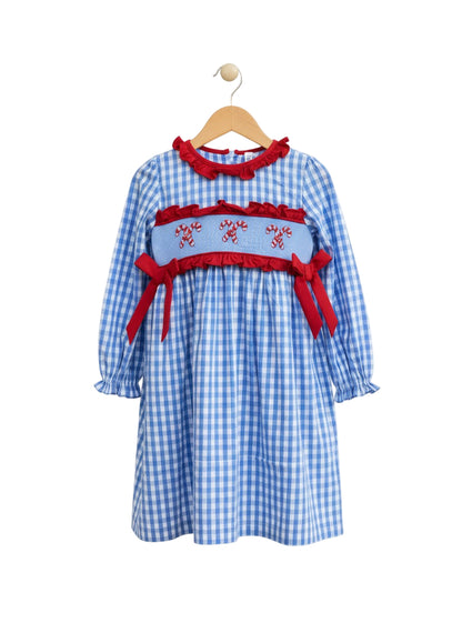 A girls' long-sleeved blue and white gingham dress hanging on a wooden hanger. It features a red ruffled collar and a smocked bodice embroidered with three pairs of candy canes, accented by red ruffles and red ribbon bows on the sides.