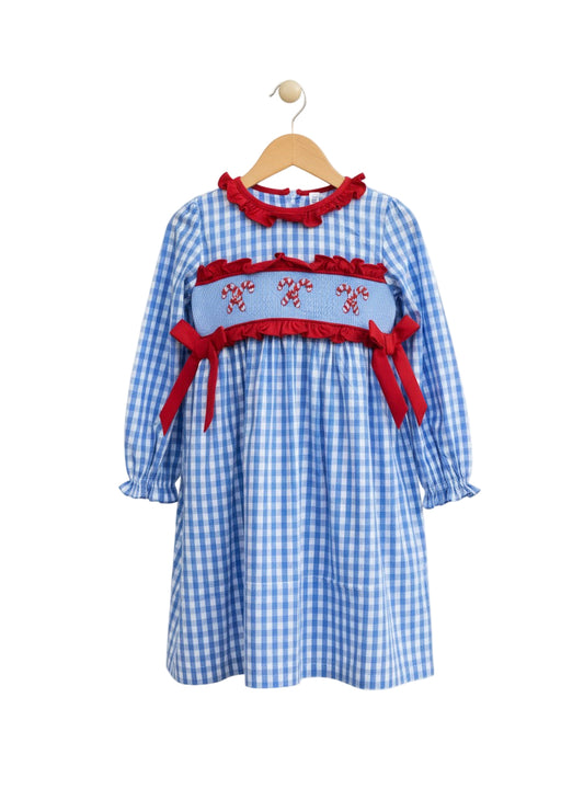 A girls' long-sleeved blue and white gingham dress hanging on a wooden hanger. It features a red ruffled collar and a smocked bodice embroidered with three pairs of candy canes, accented by red ruffles and red ribbon bows on the sides.