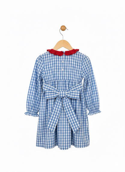 A back view of a blue and white gingham check dress for a child, hanging on a wooden hanger against a plain white background. The dress features a red ruffled collar, long sleeves with ruffled cuffs, a gathered waist, and a large matching gingham bow tied at the back. The back bodice has a button closure.
