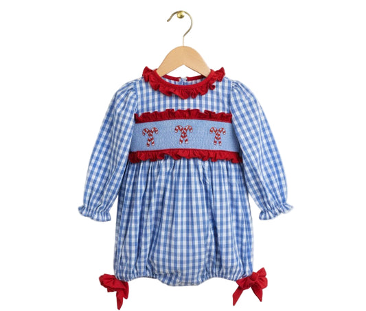 A baby girl's long-sleeved romper made of blue and white gingham fabric, hanging on a wooden hanger against a white background. It features a light blue smocked panel across the chest, framed by red ruffles, with three pairs of embroidered red and white candy canes. The long sleeves have ruffled cuffs, and the bubble-style legs are finished with large red bows at the openings.