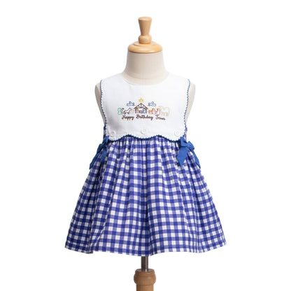 A sleeveless dress displayed on a mannequin. The dress features a white bodice with embroidered nativity scene details and the phrase ‘Happy Birthday Jesus.’ The skirt is blue and white gingham with two blue ribbon bows at the waist.