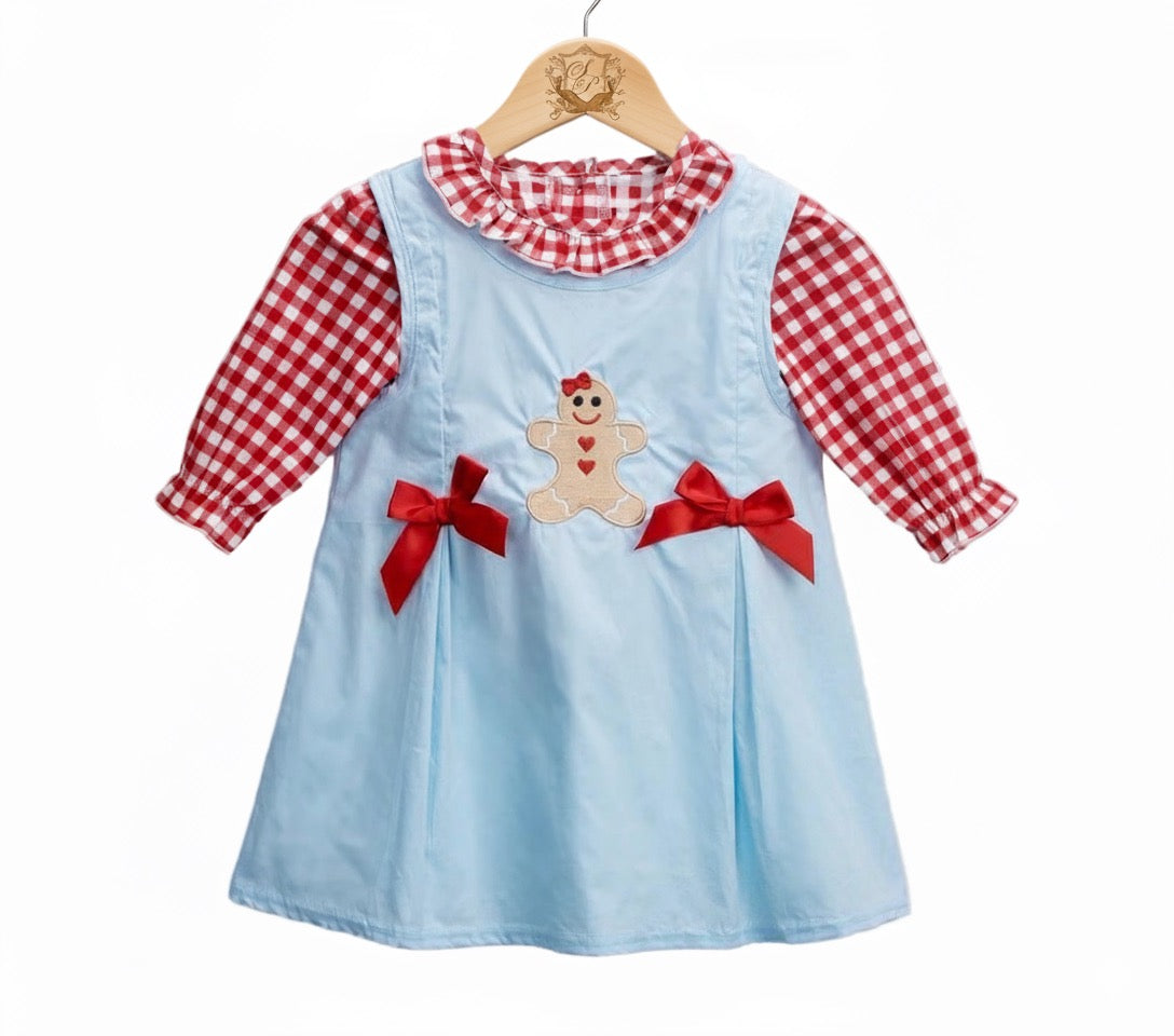 A festive baby girl outfit features a light blue jumper dress with a gingerbread girl applique and red bows, paired with a red and white gingham long-sleeve shirt with a ruffled collar, all hanging on a wooden hanger against a white background.