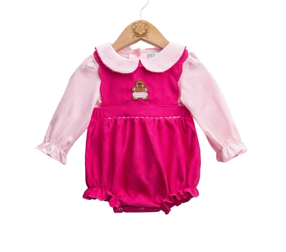 A photograph of a baby outfit hanging on a wooden hanger against a white background. The outfit consists of a light pink long-sleeved shirt with a rounded white collar and ruffled cuffs, worn underneath a bright pink corduroy bubble jumper with a gathered waist, ruffled leg openings, and an embroidered brown gingerbread man figure in the center of the chest.