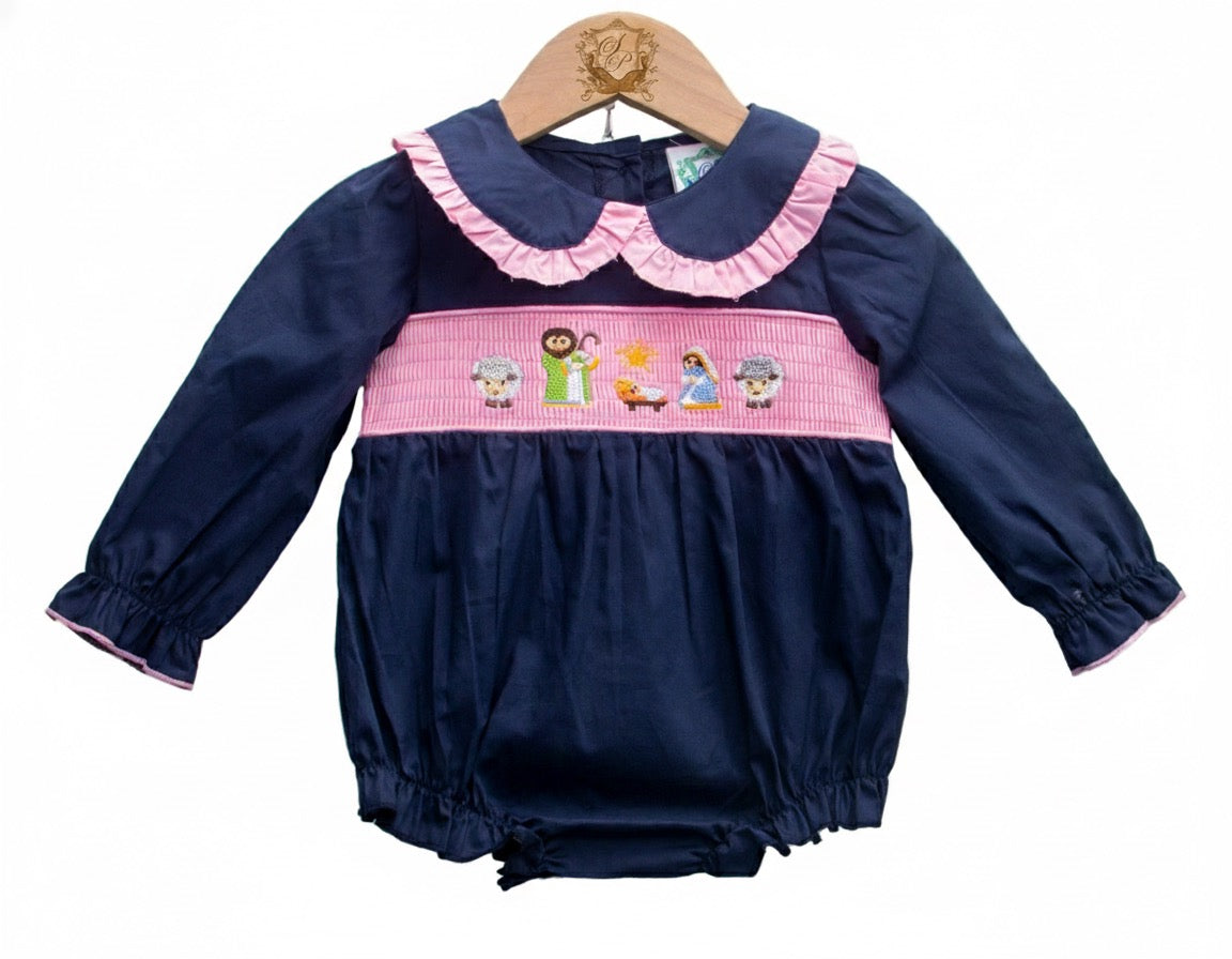 A baby romper displayed on a wooden hanger. The romper is navy blue with long sleeves and pink trim at the cuffs. It features a pink ruffled collar and a smocked waistband embroidered with nativity-themed characters, including figures and animals. The bottom has elastic leg openings.