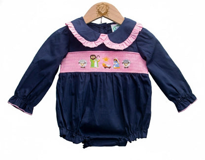 A baby romper displayed on a wooden hanger. The romper is navy blue with long sleeves and pink trim at the cuffs. It features a pink ruffled collar and a smocked waistband embroidered with nativity-themed characters, including figures and animals. The bottom has elastic leg openings.