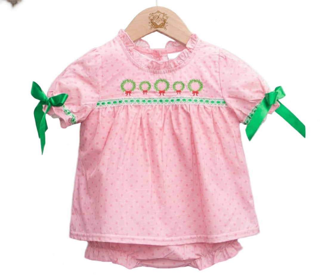 A baby diaper set displayed on a wooden hanger. The set includes a short-sleeve pink top with subtle polka dots, a ruffled neckline, and green wreath embroidery with red bows across the chest. Green ribbon bows are tied at the sleeves, and matching pink diaper cover is visible underneath.