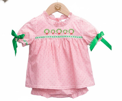 A baby diaper set displayed on a wooden hanger. The set includes a short-sleeve pink top with subtle polka dots, a ruffled neckline, and green wreath embroidery with red bows across the chest. Green ribbon bows are tied at the sleeves, and matching pink diaper cover is visible underneath.