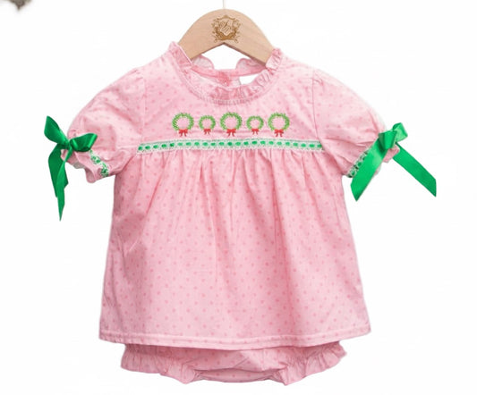 A baby diaper set displayed on a wooden hanger. The set includes a short-sleeve pink top with subtle polka dots, a ruffled neckline, and green wreath embroidery with red bows across the chest. Green ribbon bows are tied at the sleeves, and matching pink diaper cover is visible underneath.