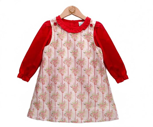A long-sleeve red dress with a sleeveless white pinafore overlay featuring a repeating pattern of pink bows and green leaves. The dress has a ruffled neckline and button details on the shoulders, displayed on a wooden hanger