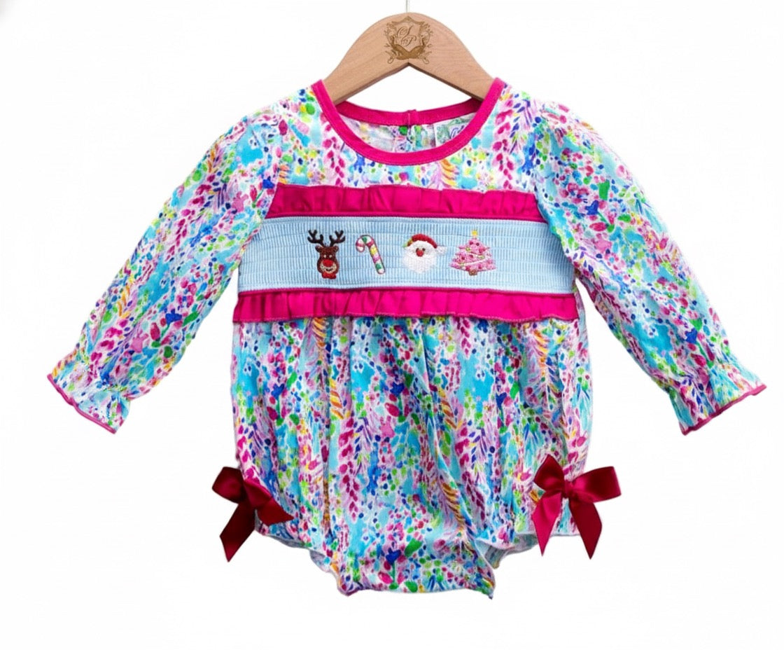 A baby girl's long-sleeved romper, featuring a colorful abstract print in blues, pinks, and greens, hangs on a wooden hanger against a white background. It has a smocked panel across the chest with embroidered Christmas motifs—a reindeer, a candy cane, Santa Claus, and a Christmas tree—framed by hot pink ruffles. The long sleeves and leg openings are trimmed in hot pink, with large red bows at the legs.