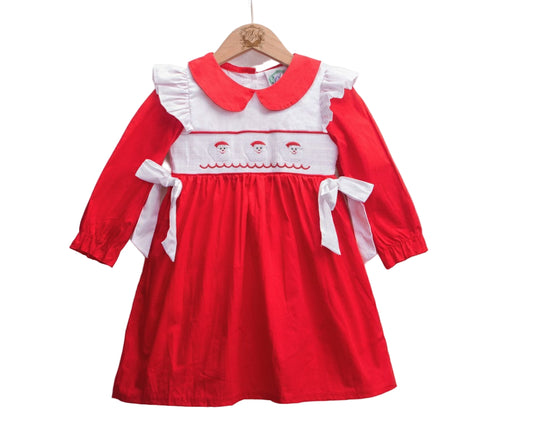 A red and white child's smocked dress hangs on a wooden hanger against a white background. The dress features a white Peter Pan collar with red trim, white ruffled shoulder accents, and a white smocked bodice with three embroidered Santa faces and a red wavy line below them. Long red sleeves end in elastic cuffs, and the red gathered skirt has a white bow tied at each side of the waist.