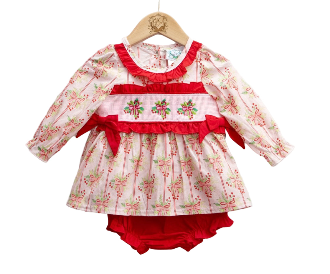 A baby diaper set displayed on a wooden hanger. The set includes a long-sleeve top with a white background and a repeating pink bow and green leaf pattern, accented by a red ruffled neckline and a smocked waistband featuring embroidered holiday bows with red ribbon details. Matching red diaper cover is visible underneath.