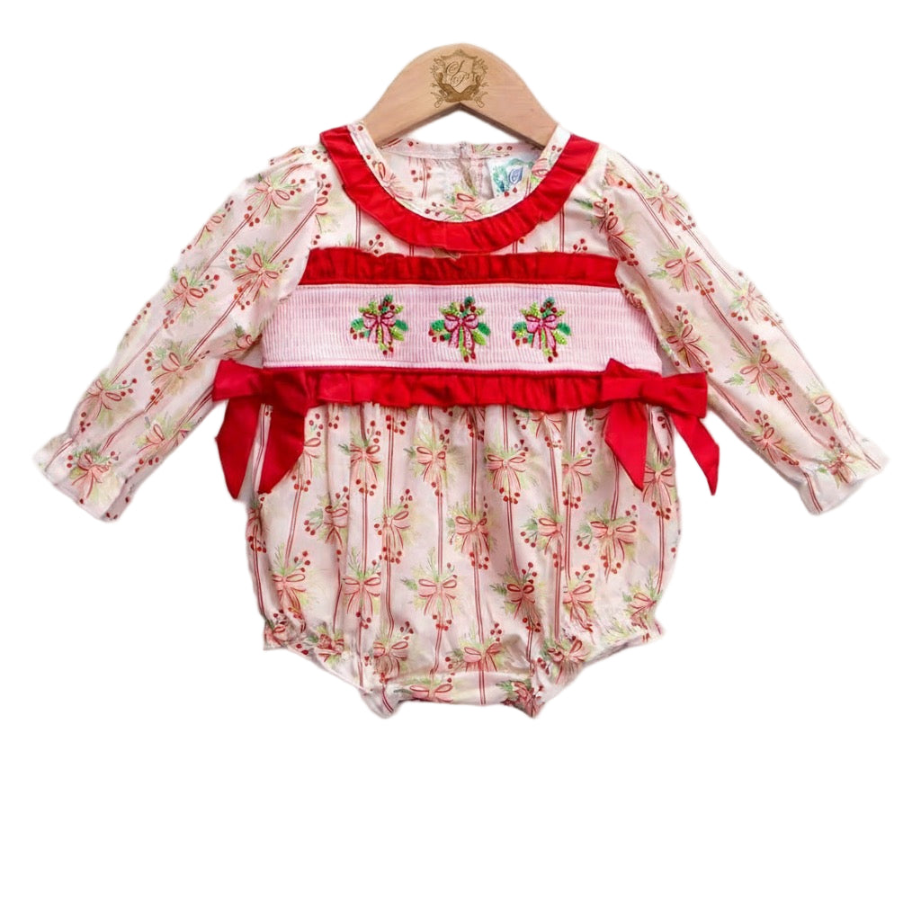 A baby romper displayed on a wooden hanger. The romper has long sleeves with a white background featuring a repeating pink bow and green leaf pattern. It includes a red ruffled neckline and a smocked waistband with embroidered holiday bows and red ribbon accents. The bottom is gathered with elastic leg openings.