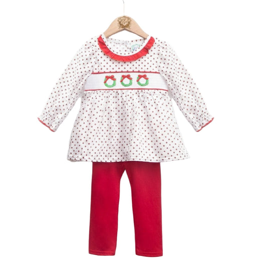 A holiday outfit displayed on a wooden hanger. The set includes a long-sleeve white tunic with small red polka dots, a ruffled red neckline, and a smocked chest panel embroidered with three green wreaths tied with red bows. The tunic is paired with solid red leggings.