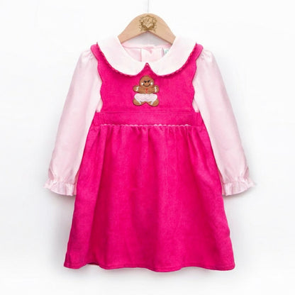A photograph shows a child's dress set hanging on a wooden hanger against a white background. The outer layer is a bright pink corduroy pinafore with a gathered skirt and a brown gingerbread man appliqué with white icing on the chest. Underneath is a light pink long-sleeved blouse with a rounded Peter Pan collar and ruffled cuffs.