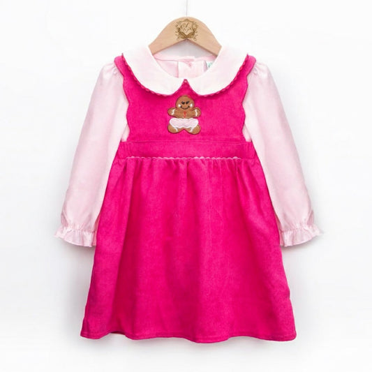A photograph shows a child's dress set hanging on a wooden hanger against a white background. The outer layer is a bright pink corduroy pinafore with a gathered skirt and a brown gingerbread man appliqué with white icing on the chest. Underneath is a light pink long-sleeved blouse with a rounded Peter Pan collar and ruffled cuffs.