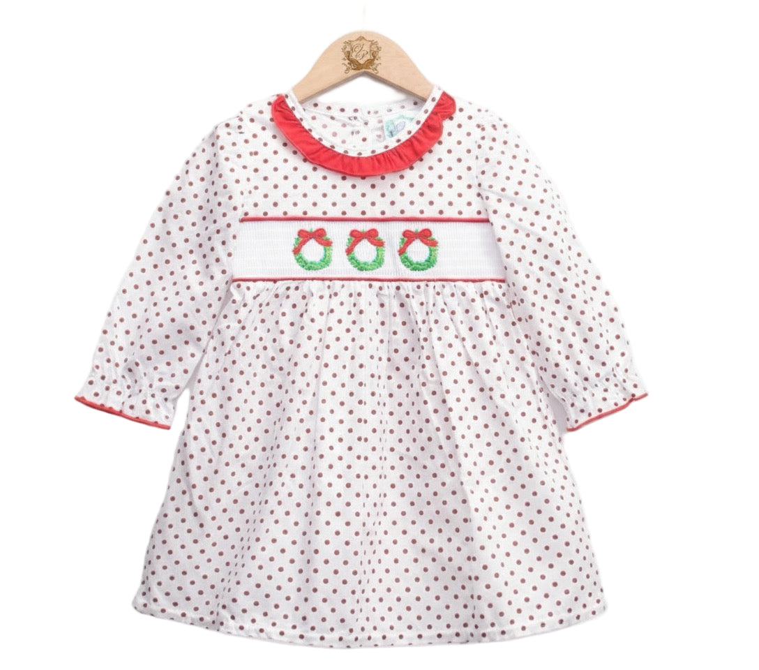 A white long-sleeved dress for a young girl featuring an all-over brown polka dot pattern, hanging on a wooden hanger. The bodice has a white smocked panel embroidered with three green Christmas wreaths topped with red bows. The neckline is accented with a red ruffled collar, and the sleeves feature matching red trim at the cuffs.