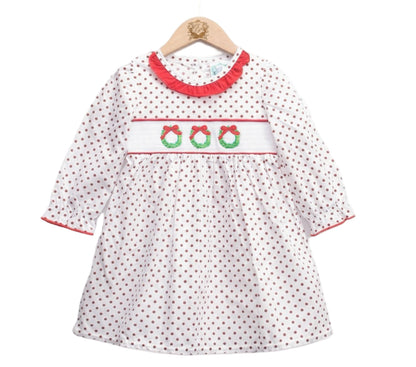 A white long-sleeved dress for a young girl featuring an all-over brown polka dot pattern, hanging on a wooden hanger. The bodice has a white smocked panel embroidered with three green Christmas wreaths topped with red bows. The neckline is accented with a red ruffled collar, and the sleeves feature matching red trim at the cuffs.