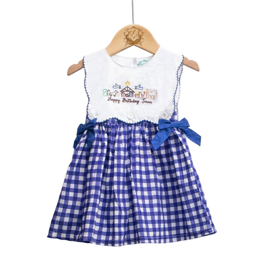 A sleeveless dress displayed on a wooden hanger. The dress features a white bodice with embroidered nativity scene details and the phrase ‘Happy Birthday Jesus.’ The skirt is blue and white gingham with two blue ribbon bows at the waist.