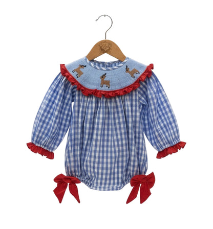 A blue and white gingham check baby romper hangs on a wooden hanger against a white background. The romper features a round, smocked yoke detailed with three embroidered brown deer and a red ruffled trim. The long sleeves have matching red ruffle cuffs. Large red velvet bows adorn the sides of the elasticized leg openings. 
