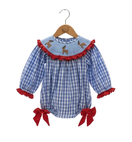 A blue and white gingham check baby romper hangs on a wooden hanger against a white background. The romper features a round, smocked yoke detailed with three embroidered brown deer and a red ruffled trim. The long sleeves have matching red ruffle cuffs. Large red velvet bows adorn the sides of the elasticized leg openings. 