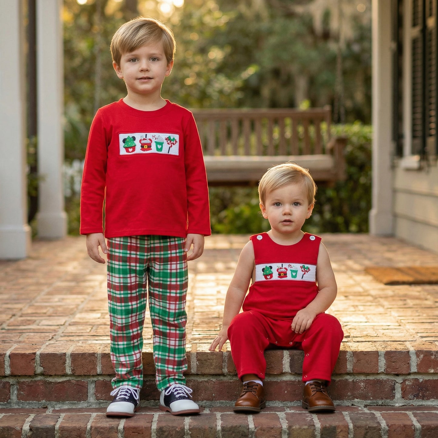 Two young boys, brothers, pose on brick steps outdoors in warm sunlight. The older boy stands on the left, wearing the red long-sleeved shirt with the smocked holiday treats panel and the green and red plaid pants from the previous image, paired with saddle shoes. The younger boy sits on the right, wearing a matching red sleeveless romper with the identical smocked panel and brown leather shoes. A wooden bench and green foliage are visible in the blurred background.