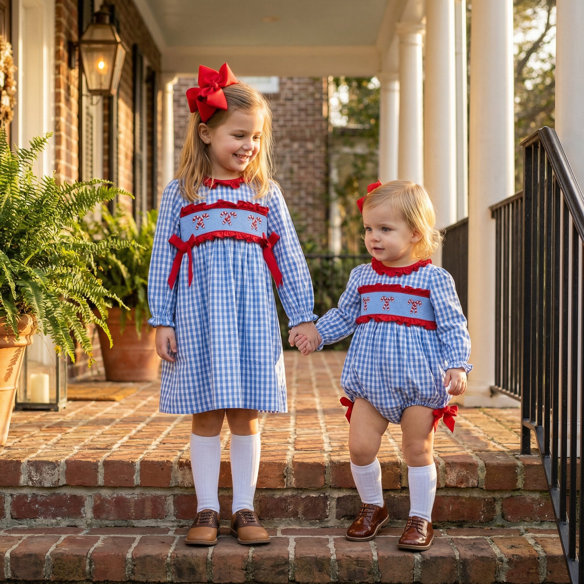 Two young sisters hold hands on brick steps, both wearing matching blue and white gingham smocked outfits with red candy cane embroidery and red bows. The older girl wears a long-sleeved dress, and the younger one wears a romper, both paired with white knee socks and brown leather shoes. A large red bow is in the older girl's hair.