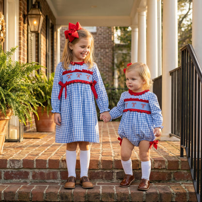 Two young sisters hold hands on brick steps, both wearing matching blue and white gingham smocked outfits with red candy cane embroidery and red bows. The older girl wears a long-sleeved dress, and the younger one wears a romper, both paired with white knee socks and brown leather shoes. A large red bow is in the older girl's hair.